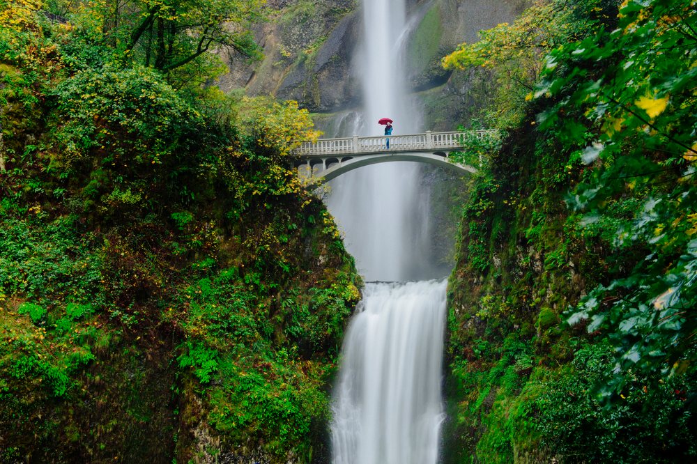 Person holding a red umbrella stands on an arched stone bridge over the multi-tiered, Multnomah Falls, Bridal Veil, Oregon.