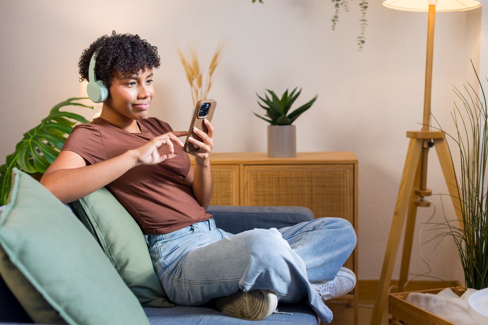 Young woman wearing wireless headphones uses smartphone while seated on a sofa in a modern living room with plants and wooden furniture.