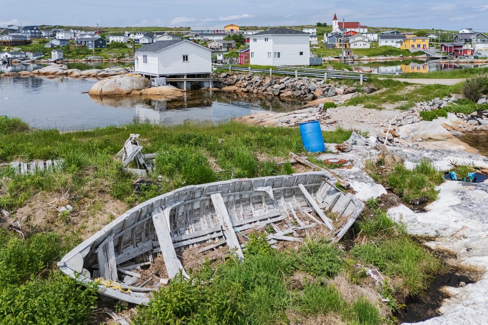 Abandoned wooden boat rests on a grassy rocky shore a small harbor with calm water and nearby houses, Landscape, Cityscape, Lifestyle, Greenspond, Newfoundland & Labrador, Canada.