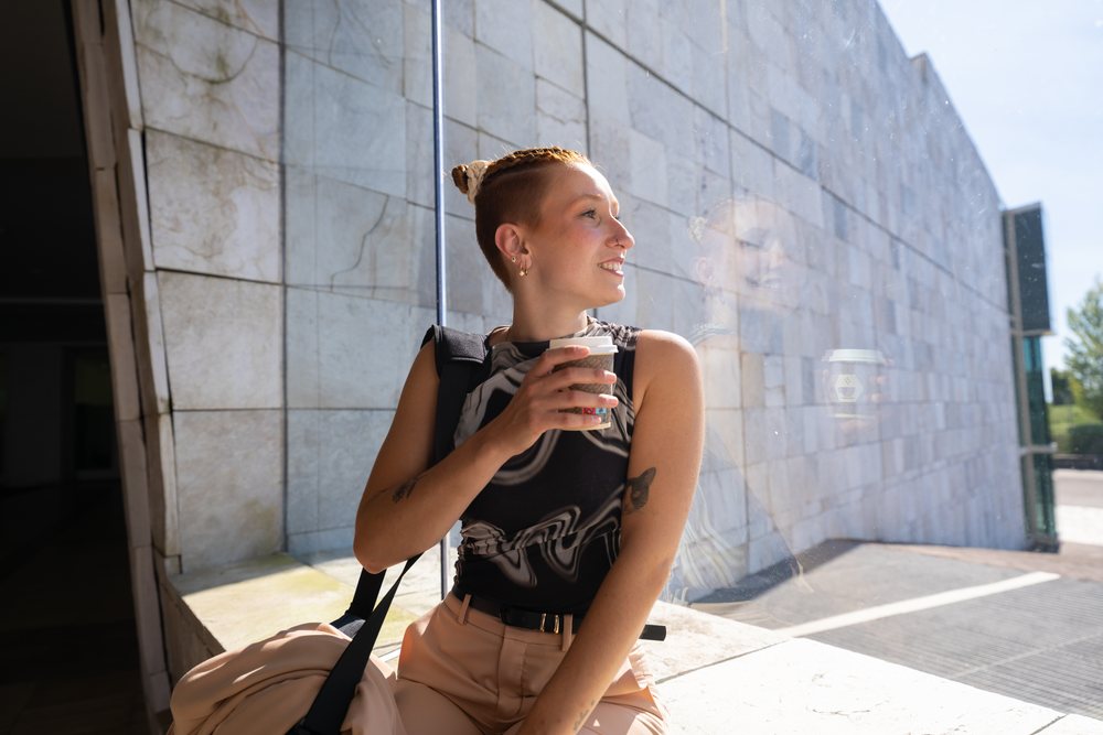 Young woman with tattoos sitting outdoors by a modern building window holding a coffee cup and smiling during daytime, businesswomen, Spain.