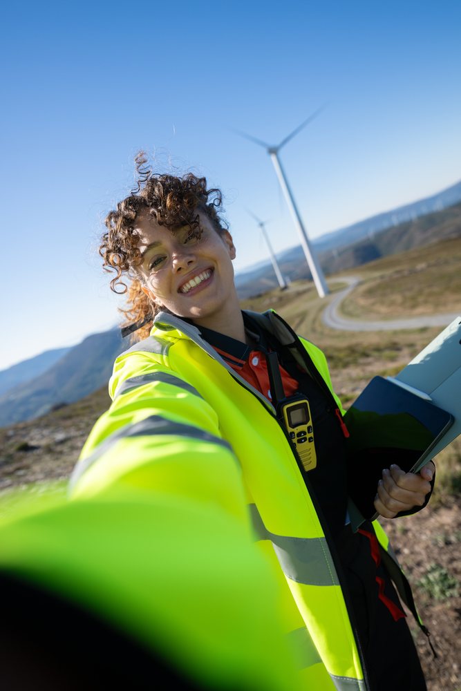 Woman wearing a high visibility jacket takes a selfie near wind turbines in rural Galicia, on a clear day, sustainability, renewable energy, Spain.