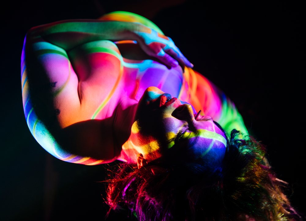 Female person with curly hair and closed eyes posing nude with rainbow colored body painting under colorful light against a dark background, pregnancy.