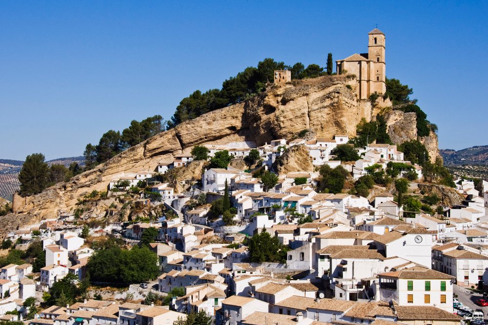 White houses cluster on a rocky hill topped by a church under a clear blue sky in Montefrio, Andalucia, Spain.