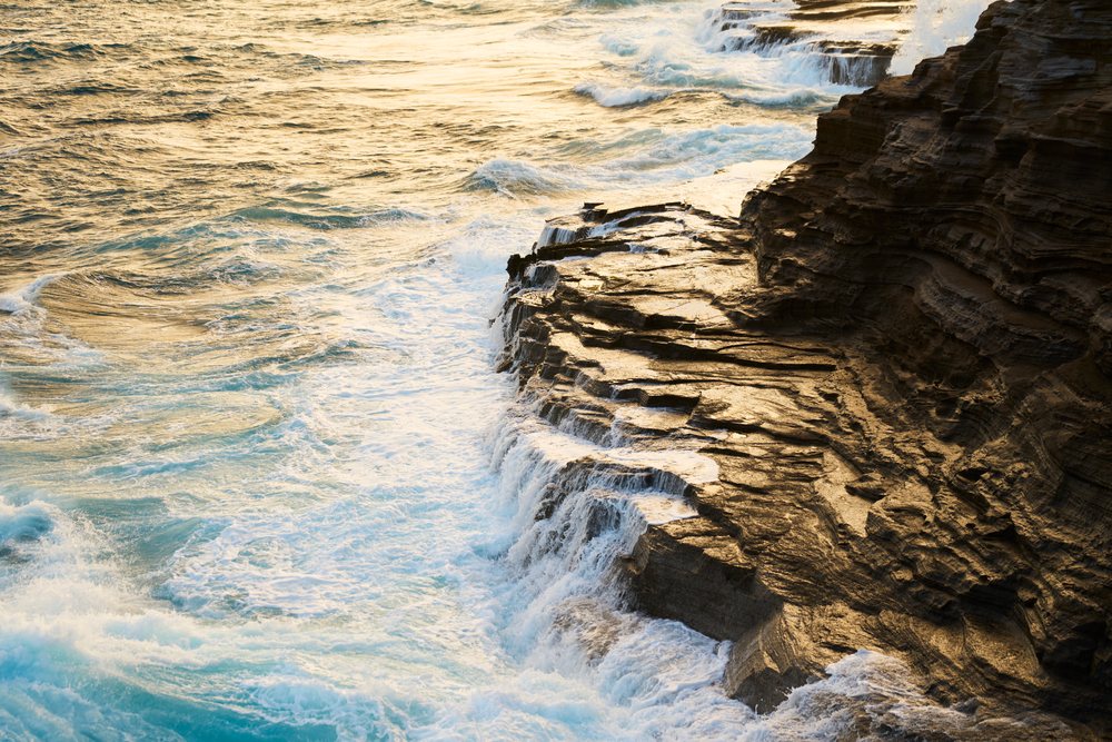 Ocean waves cascading over a rocky shoreline with sea foam at sunset, Oahu, Hawaii, USA.