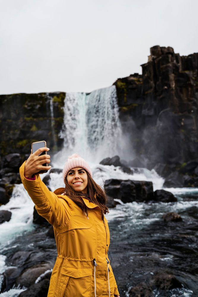A woman wearing a yellow jacket and pink beanie takes a selfie near a waterfall, Iceland.