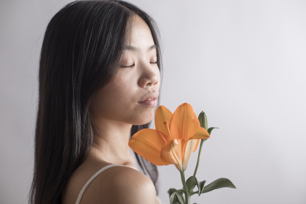 Woman with long black hair holding an orange lily near her face with closed eyes in a serene studio portrait, Art, emotional.