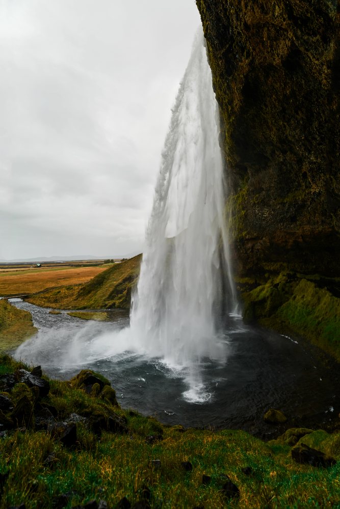 A waterfall cascades down a mossy cliff into a river pool in the Iceland countryside under an overcast sky.