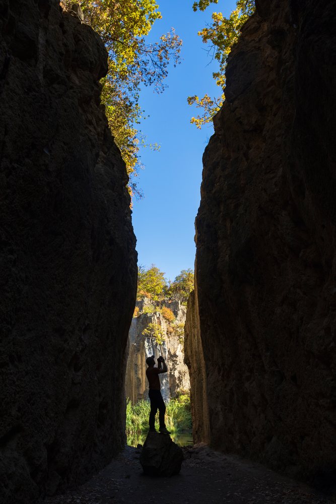 Silhouette of a photographer standing inside a narrow rocky quarry, under a blue sky with surrounding rock walls and trees above, Hungary.