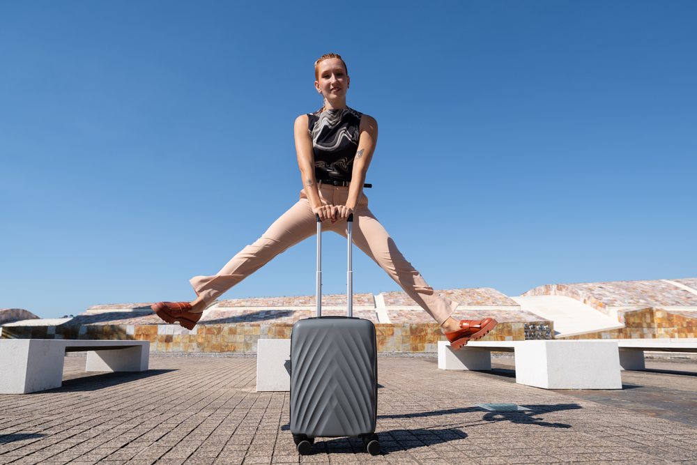 Woman jumping outdoors on a bright sunny day in Spain holding a gray hard-shell suitcase with both hands beside concrete benches and modern curved architecture, businesswomen.