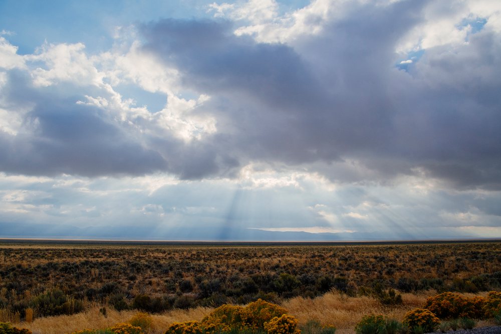 Sunrays shine through dense clouds over a dry desert landscape with shrubs and distant mountains, Nevada, USA.