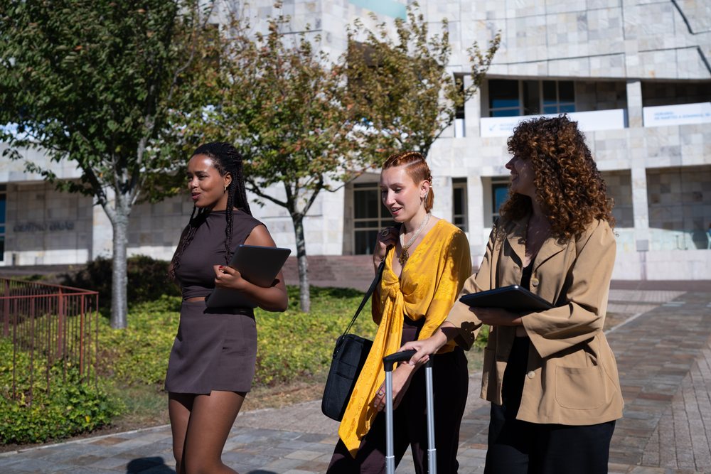 Three women walking outdoors near a modern building, two tablets and one pulling a suitcase, Spain.