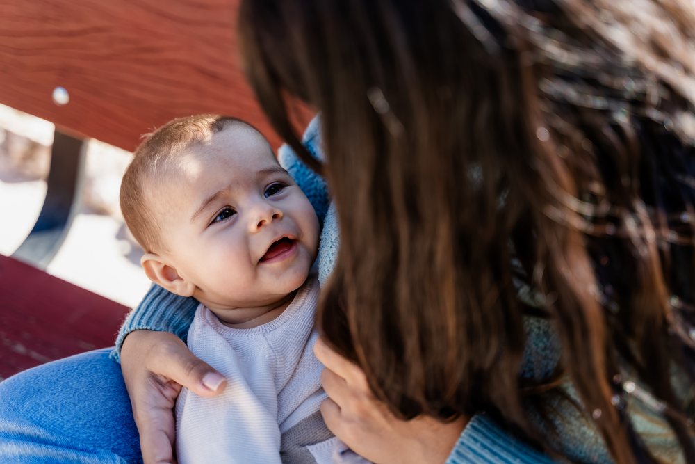 Baby smiling while held by a woman outdoors on a bench, kids, Spain.