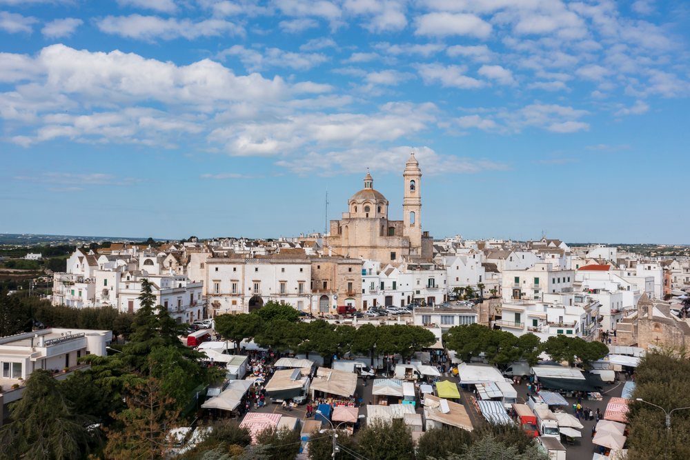 Market stalls with canopies in front of whitewashed buildings and a church tower and dome, Italy under a partly cloudy sky, Locorotondo, Puglia.