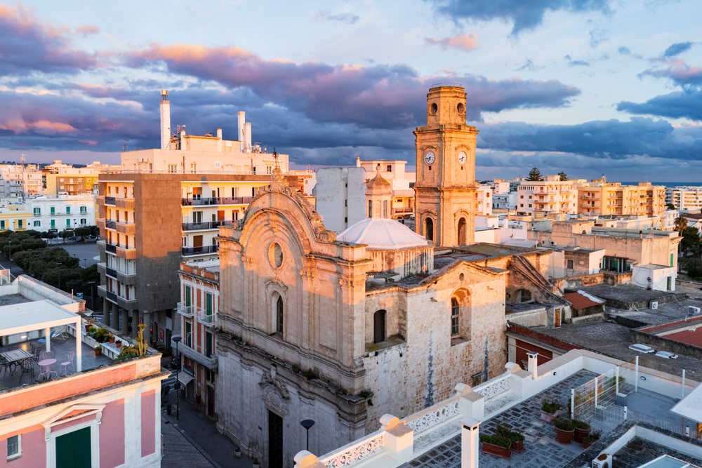 Church clock tower illuminated by sunrise light over rooftops, Monopoli, Puglia, Italy.