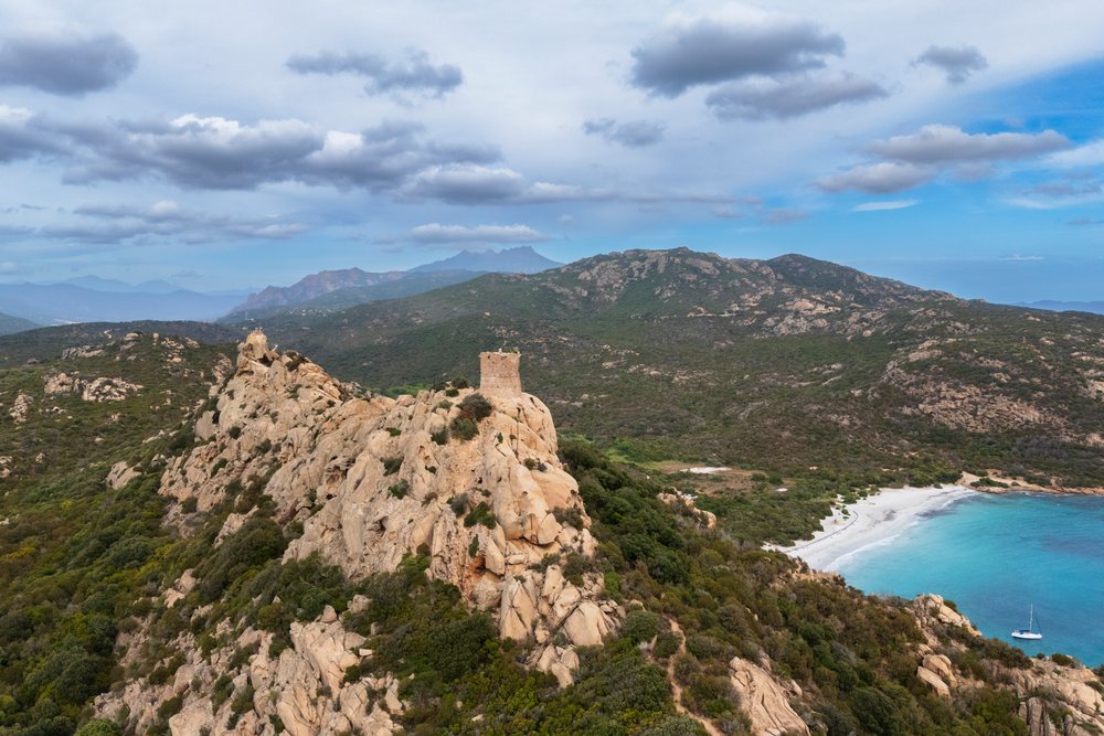 Ancient Genoese Tower stands atop a rocky hill by Cala di Roccapina beach with turquoise Mediterranean waters and Corsica mountains beyond, Travel Photography, France.