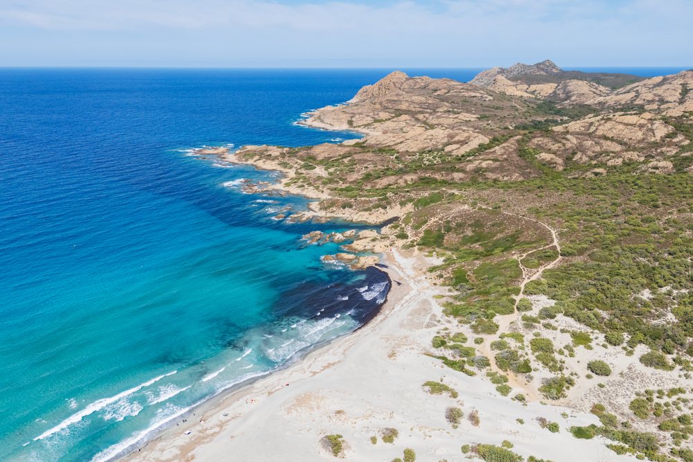 Aerial view of Plage de l'Ostriconi beach showing sandy shore, rocky coastline, Mediterranean Sea, and scattered vegetation under a clear sky, Travel Photography, Ile Rousse, Corsica, France.