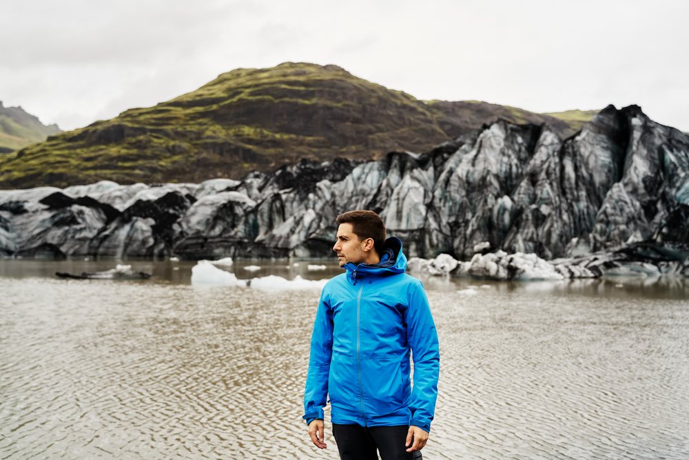 A white man stands by a glacier lake with mountains
