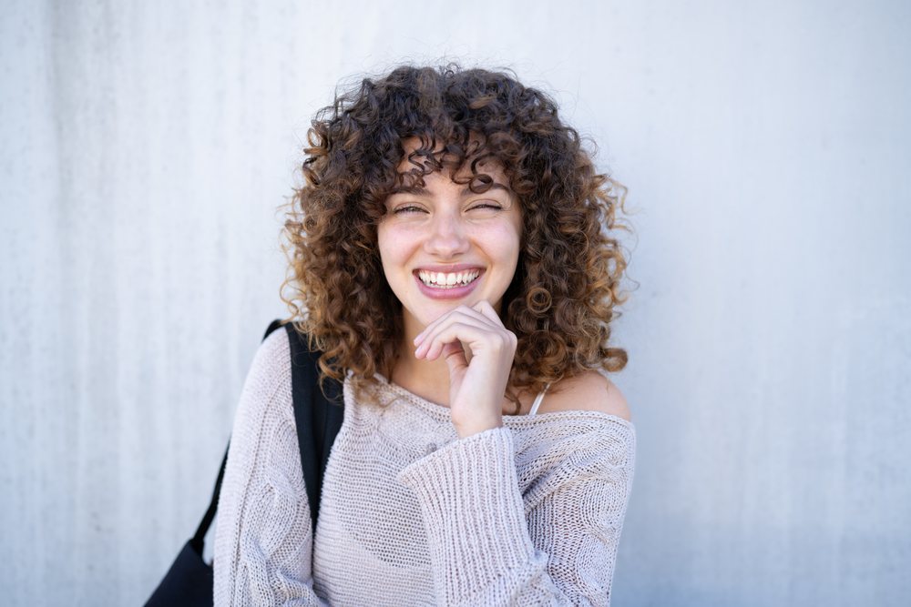 Young woman with curly hair smiling outdoors in a beige off-shoulder sweater and black shoulder bag against a light background, home story, influencer, Spain.