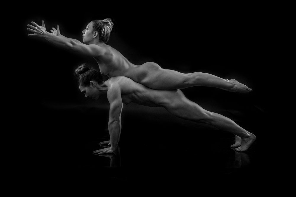 Two nude women performing partner acrobatics yoga plank pose balancing against a black background, body positivity.