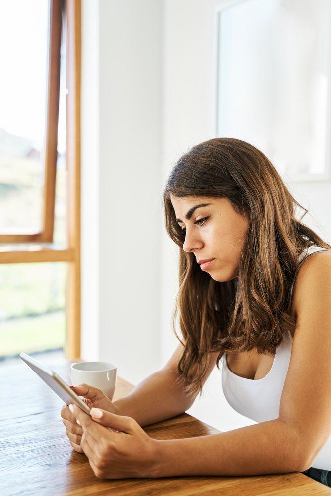 A white woman sitting at a wooden table using a tablet in a bright