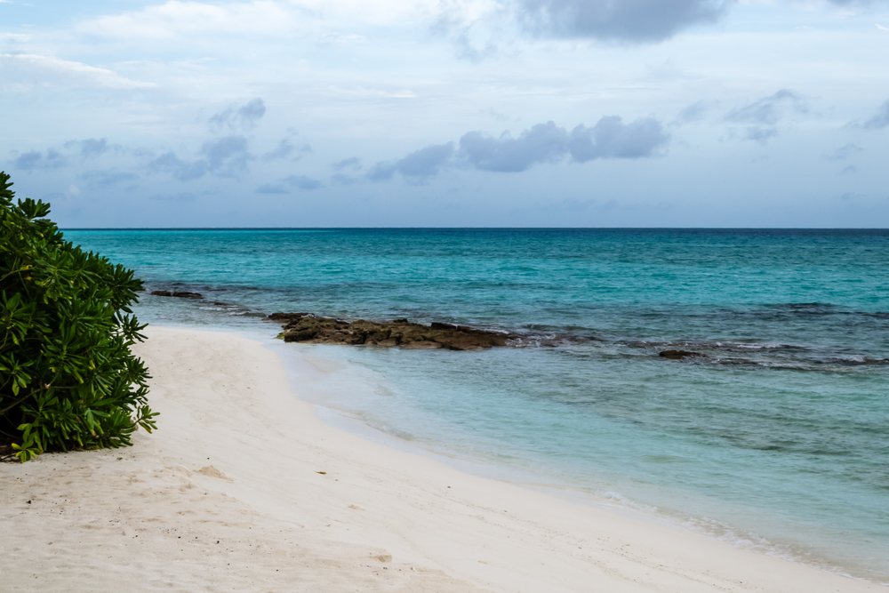 White sand beach with rocky outcrops and green foliage on the shoreline beside clear turquoise sea water under a cloudy sky, summer, Holliday, travel, landscape, Maldives.