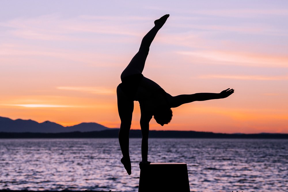 Silhouette of a woman performing a handstand on a pedestal by a lake at sunset with mountains in the background, USA.