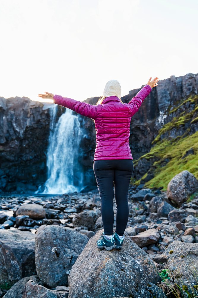 A woman stands on a rock with arms outstretched facing a waterfall, Iceland.