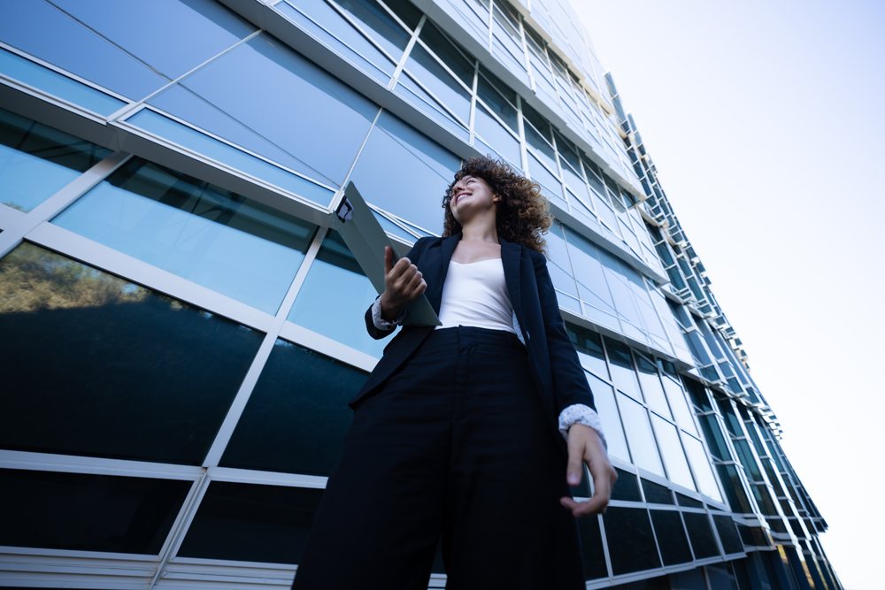 Businesswoman in a blue suit holding a folder stands outside a modern glass office building, business trip, Spain, Santiago De Compostela.