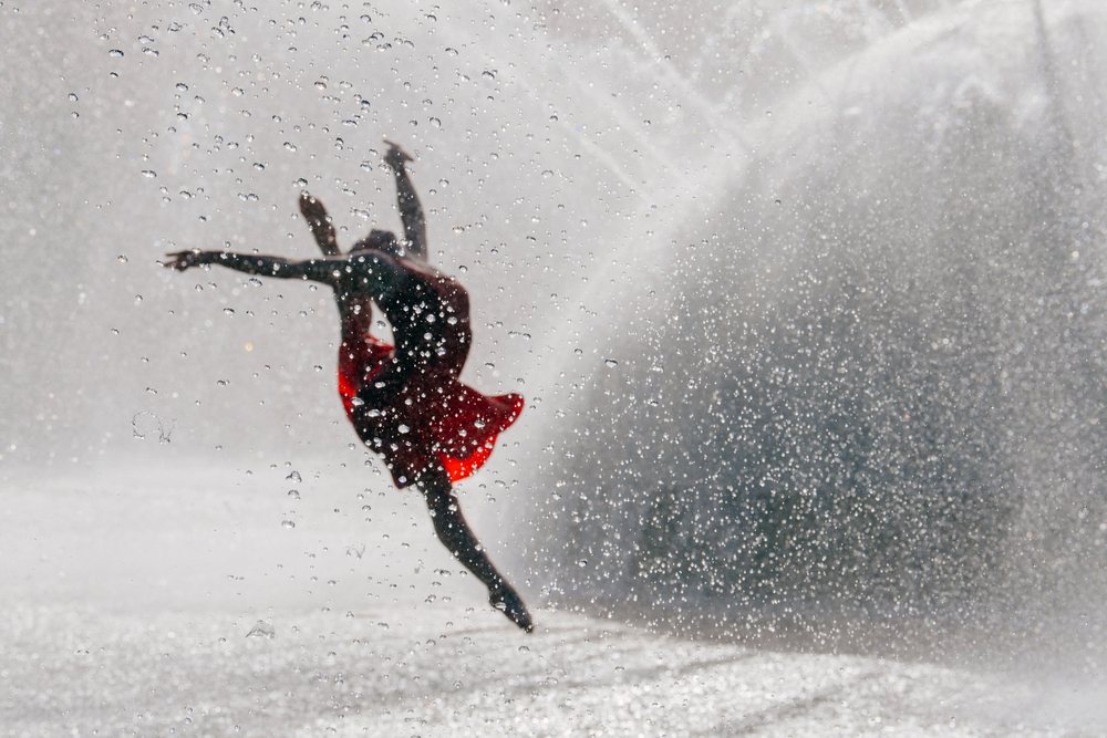 Dancer in a flowing red dress leaps gracefully through water droplets in an outdoor performance, USA.