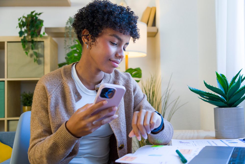 Woman in a comfortable home office sitting at a table holding a smartphone and checking her watch, remote work.