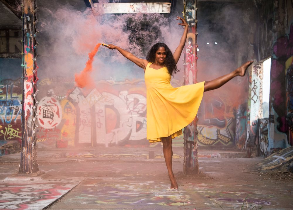 Barefoot woman dances in a yellow dress inside a graffiti-covered industrial building holding an orange smoke flare, USA.