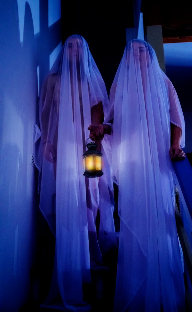 Two people dressed as ghosts with white veils hold a glowing lantern on a dimly lit staircase in darkness, USA.
