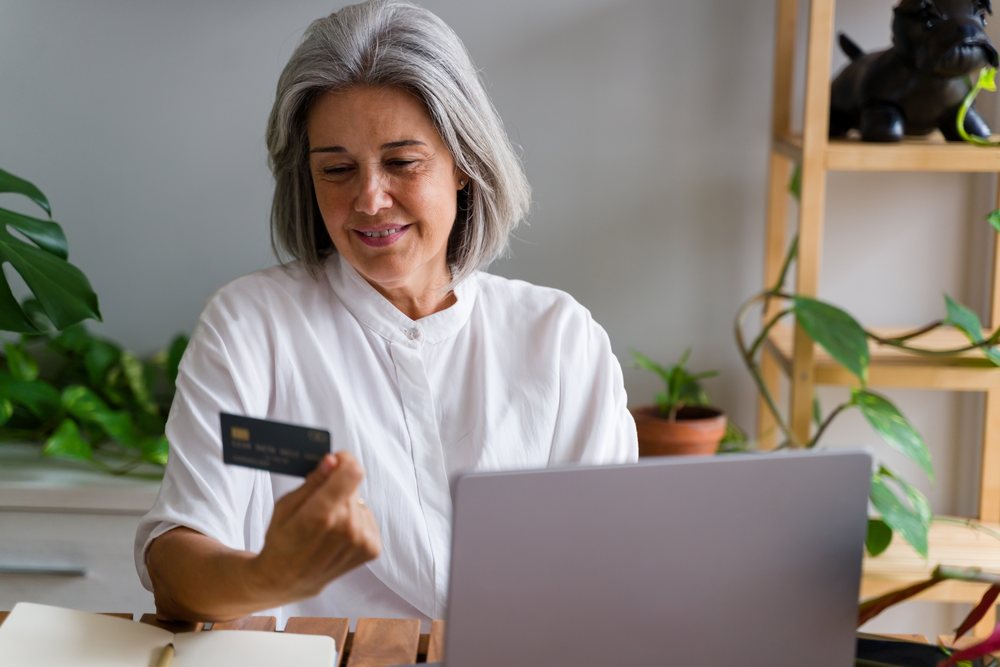 Mature woman holding a payment card and using a laptop for online shopping in a home office with plants, Spain.