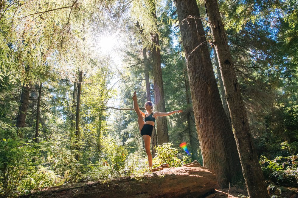 Contortionist balances on a fallen tree trunk while stretching in a sunlit forest, Tacoma, WA, USA.