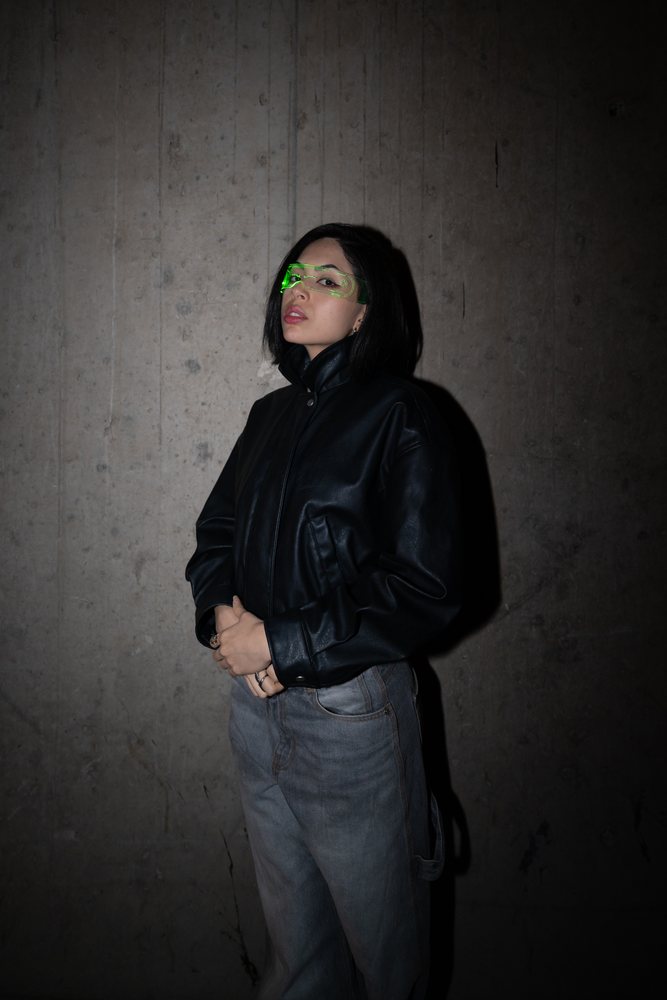Woman with short black hair stands against a concrete wall wearing a black leather jacket, blue jeans, and green transparent visor glasses in an urban style setting, Madrid, Spain.