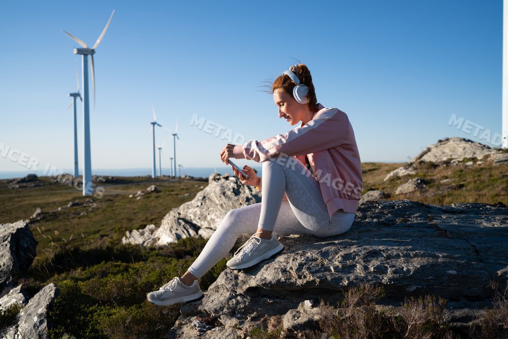 A white woman listens to music outdoors near wind turbines in Galicia