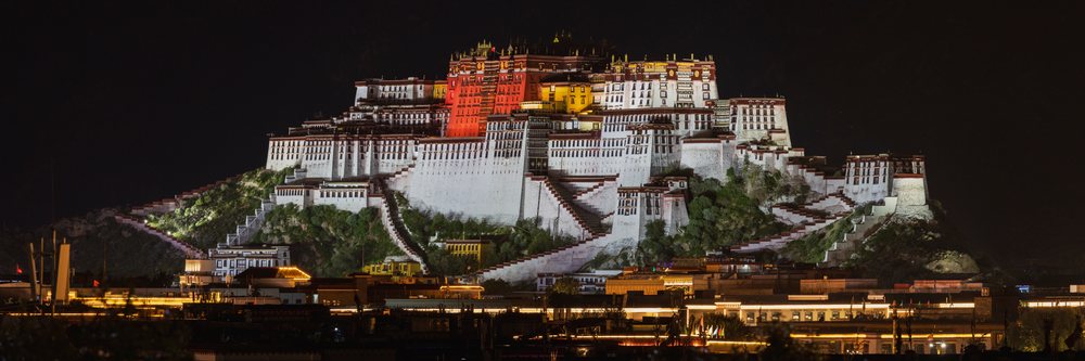 Potala Palace illuminated at Night atop a hill, Kham Tibet, displaying historic architecture and roof decorations with cityscape below, Lhasa, Autonomous Region of Tibet.