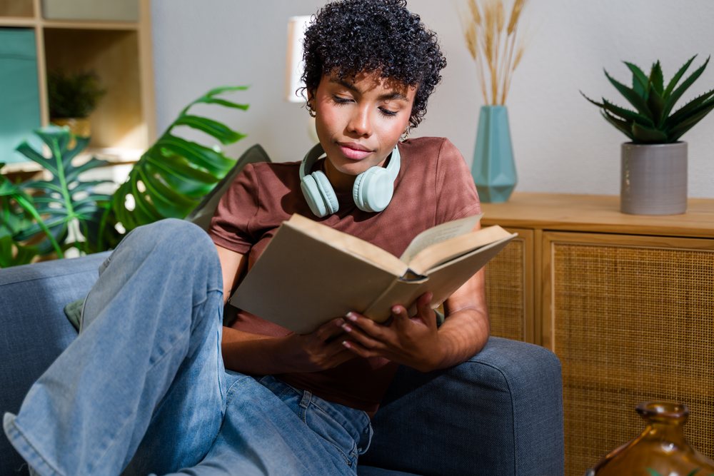 Woman seated on blue sofa reading a book with headphones around her neck in an indoor living room with plants and decorative vases, Spain.