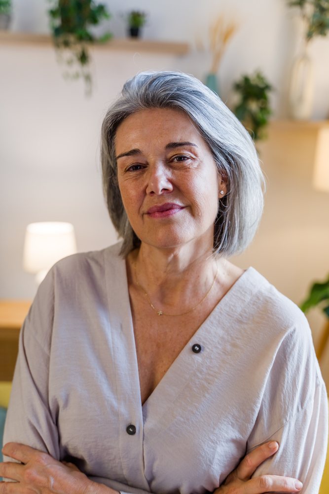 Mature woman with gray hair poses indoors at home with houseplants and a table lamp in the background, Spain.