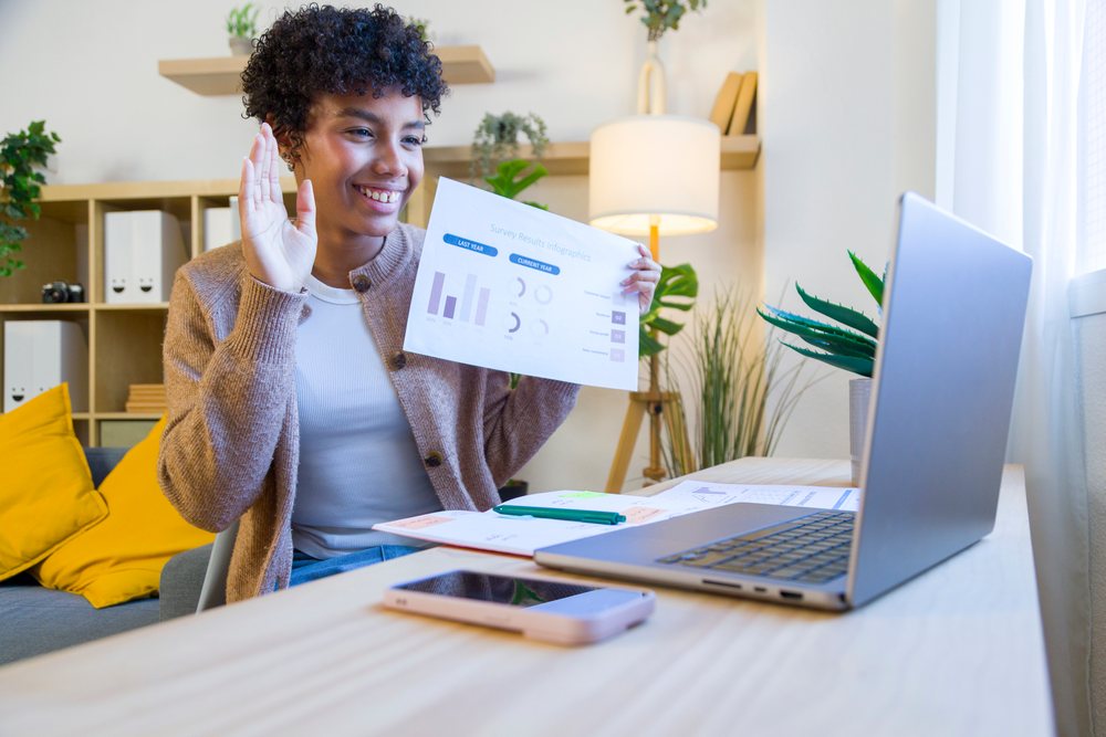 Woman in casual wear waves while holding a paper with charts during a remote video call at her home office desk.