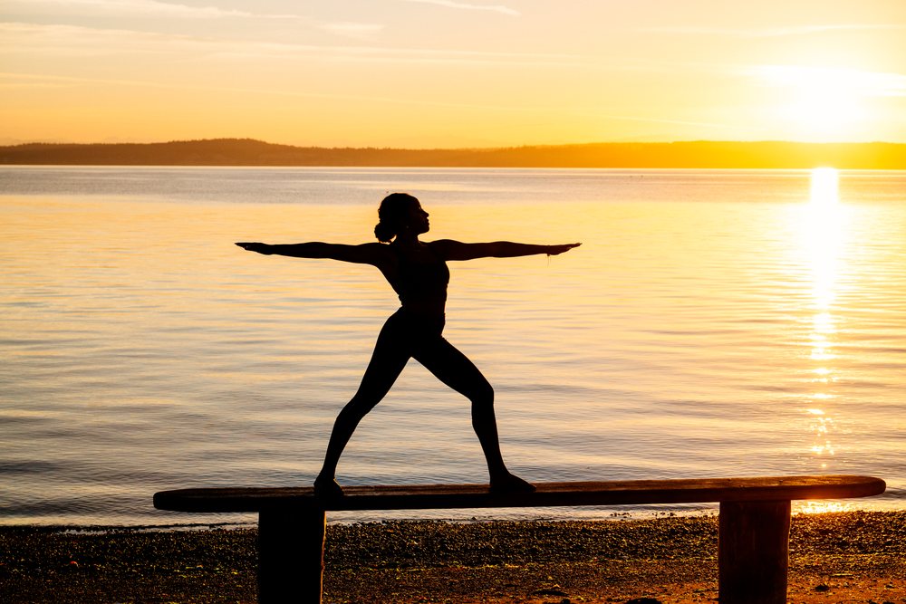 Woman practicing a yoga warrior pose balancing on a bench by a calm lake at sunset, USA.