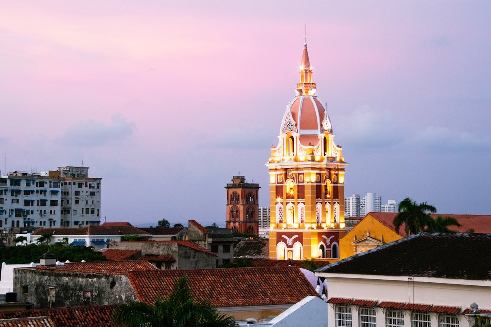 Cartagena Cathedral tower illuminated at dusk among historic city rooftops and palm trees, Colombia.