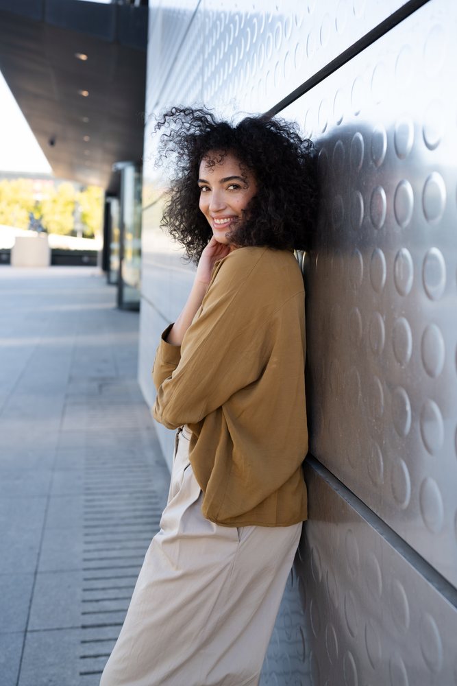 Woman with curly hair smiling outdoors while leaning against a modern textured wall, Madrid, Spain.