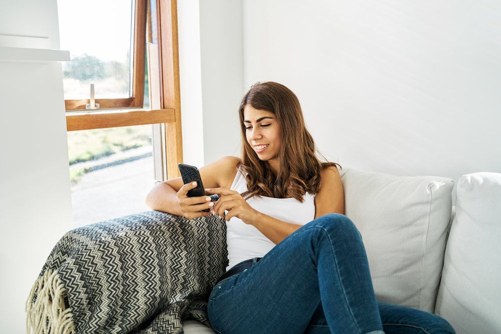A white woman relaxing on a couch and using her smartphone indoors