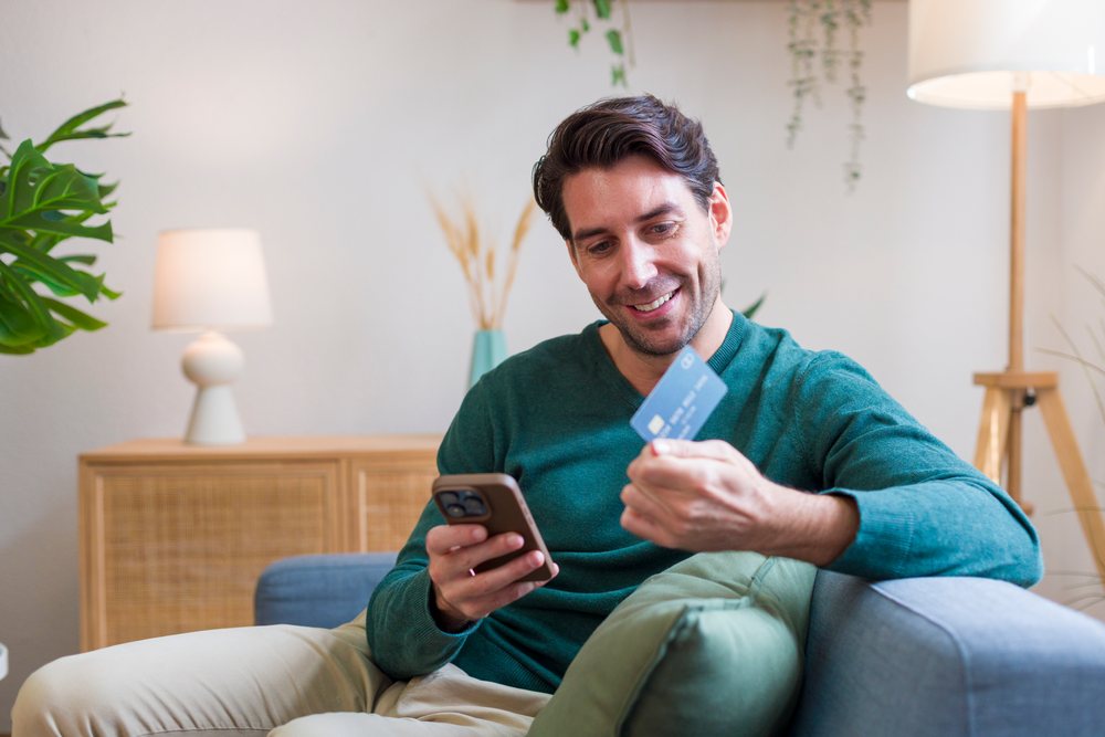 Smiling man sits on a sofa in a modern living room, using a smartphone and holding a credit card, technology, young adult, home.