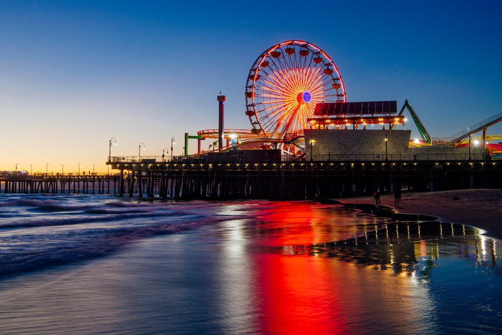 Santa Monica Pier ferris wheel illuminated at sunset reflects vibrantly on wet sand along the California  coast, CA, USA.