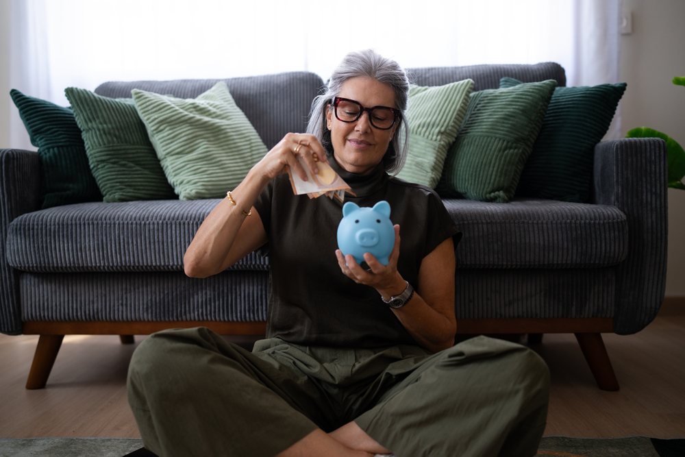 Woman with gray hair wearing eyeglasses and casual clothes sits cross-legged on the floor placing banknotes into a blue piggy bank in a living room, Spain.