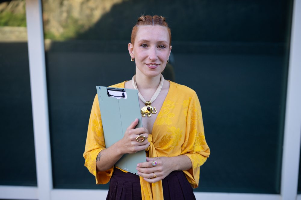 Woman with red braided hair wearing a yellow top holds a clipboard outdoors, Spain.