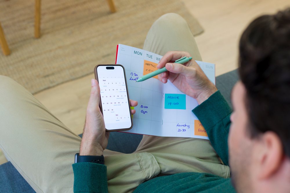 Person seated at home writing appointments on a weekly planner calendar while holding a smartphone showing a digital calendar app with sticky notes, Young adult.