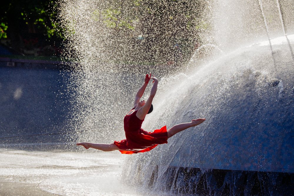 Dancer in red dress leaps midair with fountain spraying water outdoors, USA.